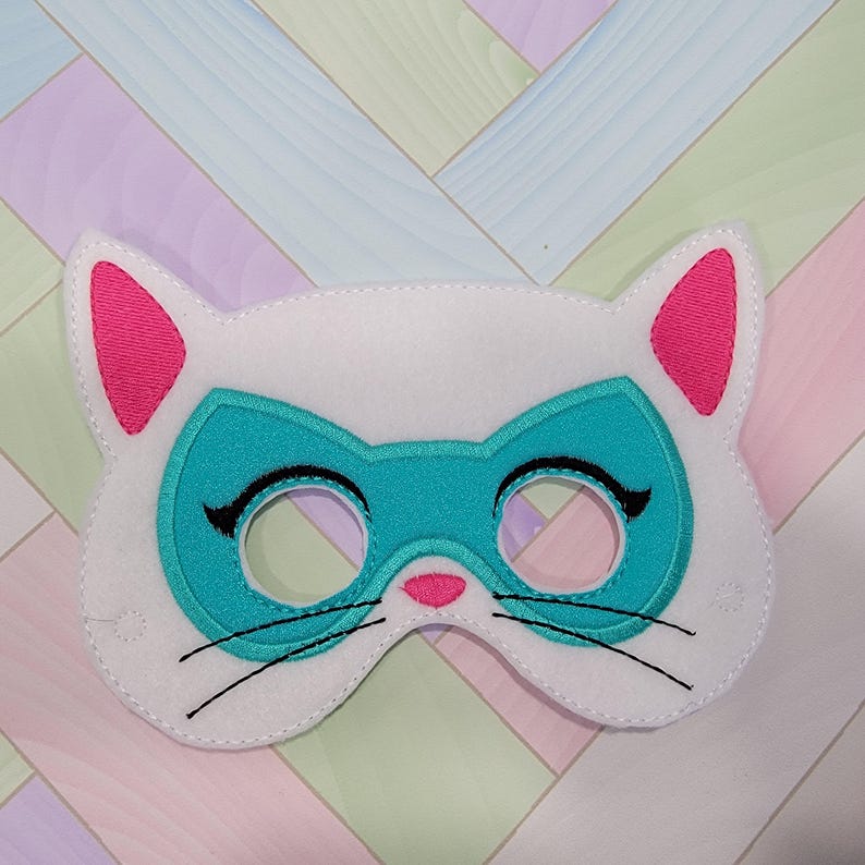 May include: A white felt cat mask featuring a teal superhero-style eye mask overlay, bright pink inner ears, a small pink nose, and black embroidered whiskers. The mask has two circular eye cutouts with black stitched eyelashes on the outer edges.