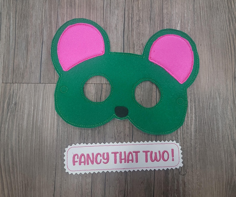 May include: A green felt animal mask with bright pink inner ears and a small black nose. Below the mask is a white rectangular felt patch with pink scalloped edges featuring the embroidered pink text "FANCY THAT TWO!" in a bold font.