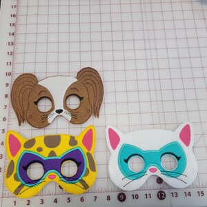 May include: Three felt animal masks featuring a brown and white dog mask with long ears, a yellow and brown leopard mask with a purple eye patch, and a white cat mask with a teal eye patch, all displayed on a grid mat with centimetre markings.