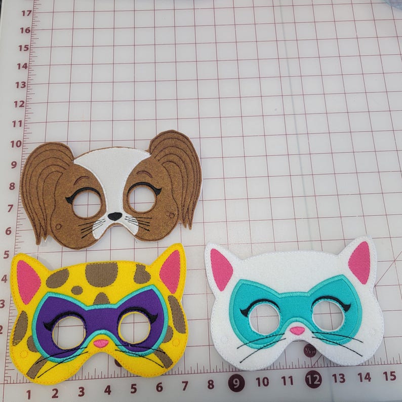 May include: Three felt animal masks featuring a brown and white dog mask with long ears, a yellow and brown leopard mask with a purple eye patch, and a white cat mask with a teal eye patch, all displayed on a grid mat with centimetre markings.