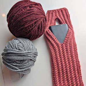 Könnte beinhalten: Ein burgunderfarbener Wollknäuel und ein grauer Wollknäuel liegen neben einer rosa gestrickten Handyhülle. Die Handyhülle hat eine dreieckige Öffnung für den Telefonbildschirm. Die Hülle ist aus Rippenstrick gefertigt.