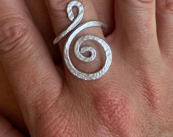May include: A silver ring with a textured, spiral design. The ring is worn on a finger.