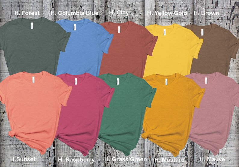 May include: Ten different colored t-shirts laid out on a wooden surface. The colors are Forest, Columbia Blue, Clay, Yellow Gold, Brown, Sunset, Raspberry, Grass Green, Mustard, and Mauve.