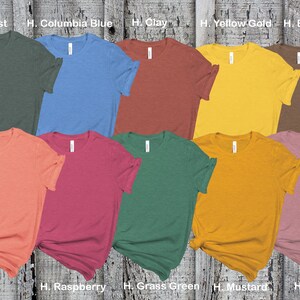 K&ouml;nnte beinhalten: Zehn T-Shirts in verschiedenen Farben, die auf einer Holzoberfl&auml;che ausgelegt sind. Die Farben sind Waldgr&uuml;n, Columbia-Blau, Ton, Goldgelb, Braun, Sonnenuntergang, Himbeere, Grasgr&uuml;n, Senf und Mauve.