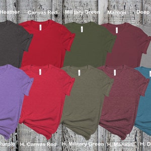 May include: Ten blank t-shirts in various colors, including dark grey heather, canvas red, military green, maroon, deep heather, team purple, canvas red, military green, maroon, and deep teal. The shirts are laid out on a wooden surface.