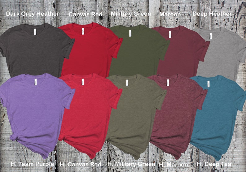 May include: Ten blank t-shirts in various colors, including dark grey heather, canvas red, military green, maroon, deep heather, team purple, canvas red, military green, maroon, and deep teal. The shirts are laid out on a wooden surface.