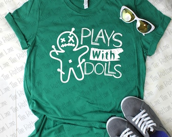 Puede incluir: Una camiseta verde con un gráfico blanco de una figura de palo con una X por ojos y el texto "PLAYS WITH DOLLS".