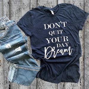 May include: A dark blue t-shirt with the text "Don't Quit Your Day Dream" in white lettering. The shirt is laying on a wooden surface with a pair of blue jeans.