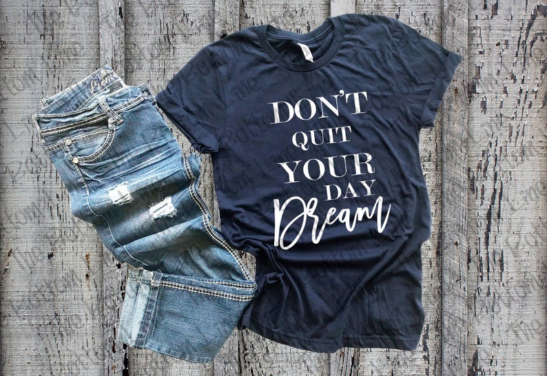 May include: A dark blue t-shirt with the text "Don't Quit Your Day Dream" in white lettering. The shirt is laying on a wooden surface with a pair of blue jeans.