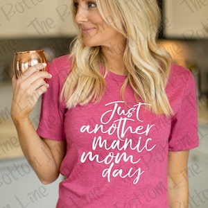May include: A woman wearing a pink t-shirt with the text "Just another manic mom day" in white lettering. She is holding a copper mug in her hand.