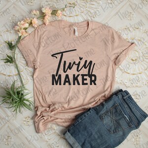 May include: A light pink t-shirt with the text "Twiy MAKER" in black lettering. The shirt is laying on a white surface with a pair of blue jeans folded next to it.