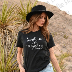 K&ouml;nnte beinhalten: Ein schwarzes T-Shirt mit wei&szlig;em Text, der "Sunshine & Whiskey" lautet, mit einem wei&szlig;en Pfeil, der nach rechts zeigt.