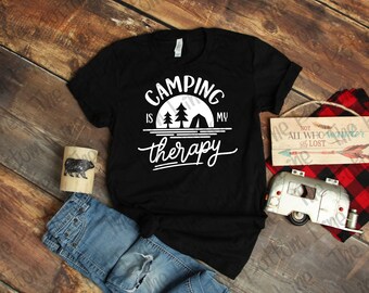 May include: Black t-shirt with the white text "Camping is my therapy" in a stylized font. The design includes a camping scene with trees and a tent. The shirt is paired with blue jeans and camping-themed decor.