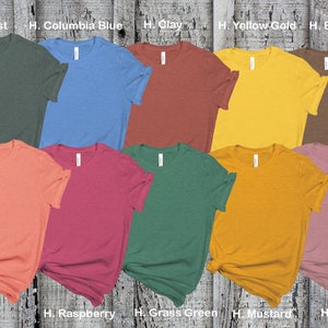 May include: Ten different colored t-shirts laid out on a wooden surface. The colors are forest green, columbia blue, clay, yellow gold, brown, sunset, raspberry, grass green, mustard, and mauve.