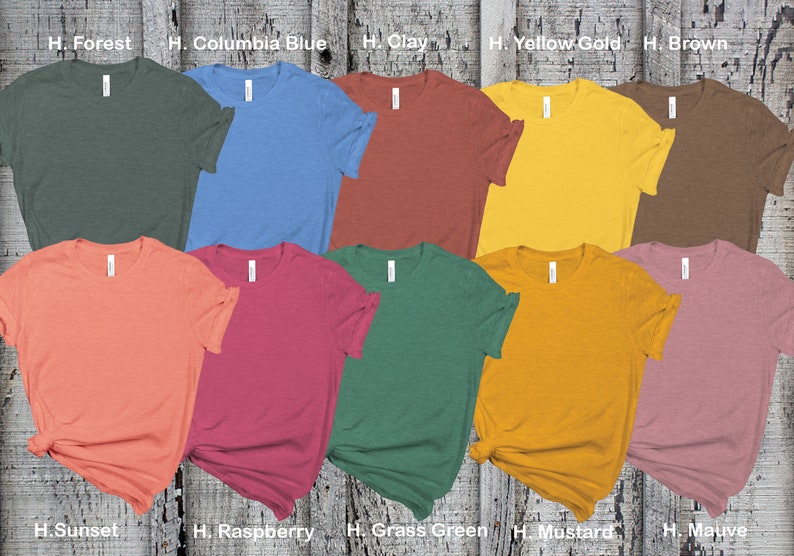 May include: Ten different colored t-shirts laid out on a wooden surface. The colors are forest green, columbia blue, clay, yellow gold, brown, sunset, raspberry, grass green, mustard, and mauve.