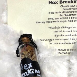 May include: A small glass bottle filled with dried herbs and flowers, sealed with black wax. A black label with the words "Hex Breaking" in white is attached to the bottle. A card with text about hex breaking is in the background.