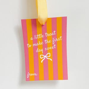 Pu&ograve; includere: Un'etichetta regalo rettangolare con strisce verticali rosa e arancioni, sospesa da un nastro giallo. L'etichetta presenta il testo "a little treat to make the first day sweet" e un'immagine di fiocco.