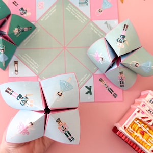 May include: A collection of paper fortune tellers and a game board with ballet and nutcracker illustrations. The fortune tellers are teal and pink, with gold star garland and a pink background. A decorative ornament is also present.