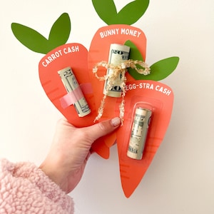 Op de afbeelding: Drie oranje wortelvormige cadeaubonnenhouders met groene bladeren. Elk bevat een opgerold biljet, met de tekst "Carrot Cash", "Bunny Money" en "Egg-stra Cash". Eén biljet is vastgezet met roze tape en een ander is vastgebonden met een gouden lint.