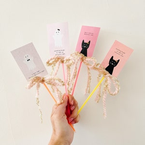 May include: Four pink and white Halloween themed cards with black and white illustrations of a ghost and a cat. The cards are attached to colorful straws with pink and gold tinsel. The straws are held in a hand.