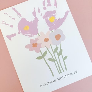 May include: A white card with two purple handprints that are painted to look like flowers. The handprints are surrounded by four pastel-colored flowers with green stems and leaves. The text "HANDMADE WITH LOVE BY" is printed at the bottom of the card.