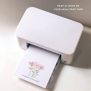 May include: A white printer with a sheet of paper coming out of it. The paper has a simple illustration of three pink and purple flowers with green stems. The text "HANDMADE WITH LOVE BY" is printed at the bottom of the page.