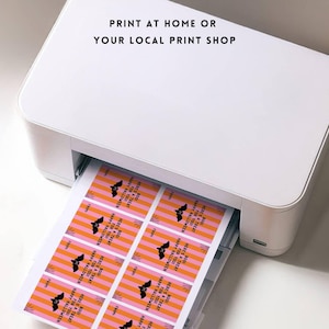 May include: A white printer printing Halloween-themed gift tags. The tags have a pink and orange striped background with a black bat graphic and the text "Happy Halloween" and "Here's a treat". The text "Print at home or your local print shop" is at the top.