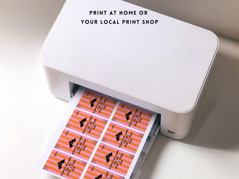 May include: A white printer printing Halloween-themed gift tags. The tags have a pink and orange striped background with a black bat graphic and the text "Happy Halloween" and "Here's a treat". The text "Print at home or your local print shop" is at the top.
