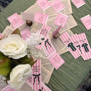 Pode incluir: Uma exibi&ccedil;&atilde;o de itens com tema de clube do livro em uma superf&iacute;cie verde. Marcadores listrados rosa e branco com la&ccedil;os verdes e texto est&atilde;o espalhados entre p&aacute;ginas de livros abertas. Pequenos cart&otilde;es rosa com perguntas relacionadas a livros tamb&eacute;m est&atilde;o presentes, junto com rosas brancas.