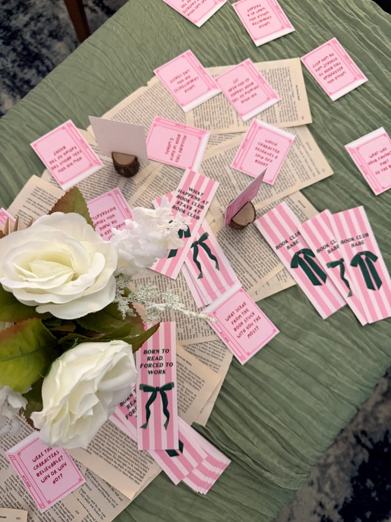 Pode incluir: Uma exibi&ccedil;&atilde;o de itens com tema de clube do livro em uma superf&iacute;cie verde. Marcadores listrados rosa e branco com la&ccedil;os verdes e texto est&atilde;o espalhados entre p&aacute;ginas de livros abertas. Pequenos cart&otilde;es rosa com perguntas relacionadas a livros tamb&eacute;m est&atilde;o presentes, junto com rosas brancas.