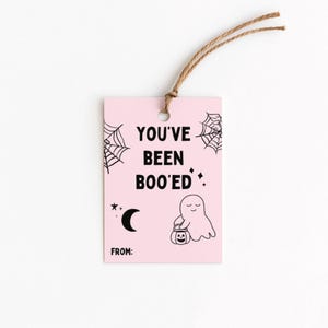 May include: A pink Halloween gift tag with the text "YOU'VE BEEN BOO'ED" in black. The tag features a ghost holding a jack-o'-lantern, spiderwebs, a crescent moon, and stars. A brown string is attached at the top.