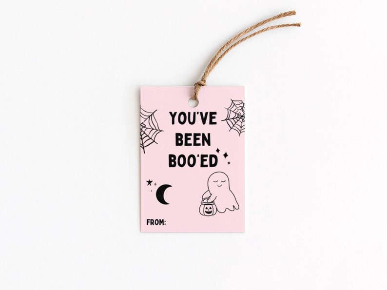 May include: A pink Halloween gift tag with the text "YOU'VE BEEN BOO'ED" in black. The tag features a ghost holding a jack-o'-lantern, spiderwebs, a crescent moon, and stars. A brown string is attached at the top.