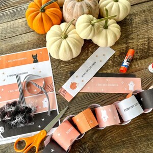 May include: A Halloween countdown chain with orange and black paper circles, each with a number and a Halloween-themed image. The chain is partially assembled on a wooden surface with white pumpkins and a glue stick.
