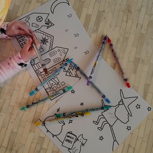 Puede incluir: Papel blanco con dibujos lineales negros de una casa embrujada, una bruja en una escoba y estrellas. Varios l&aacute;pices de colores est&aacute;n esparcidos alrededor de los dibujos. Una persona sostiene un bol&iacute;grafo sobre el dibujo.