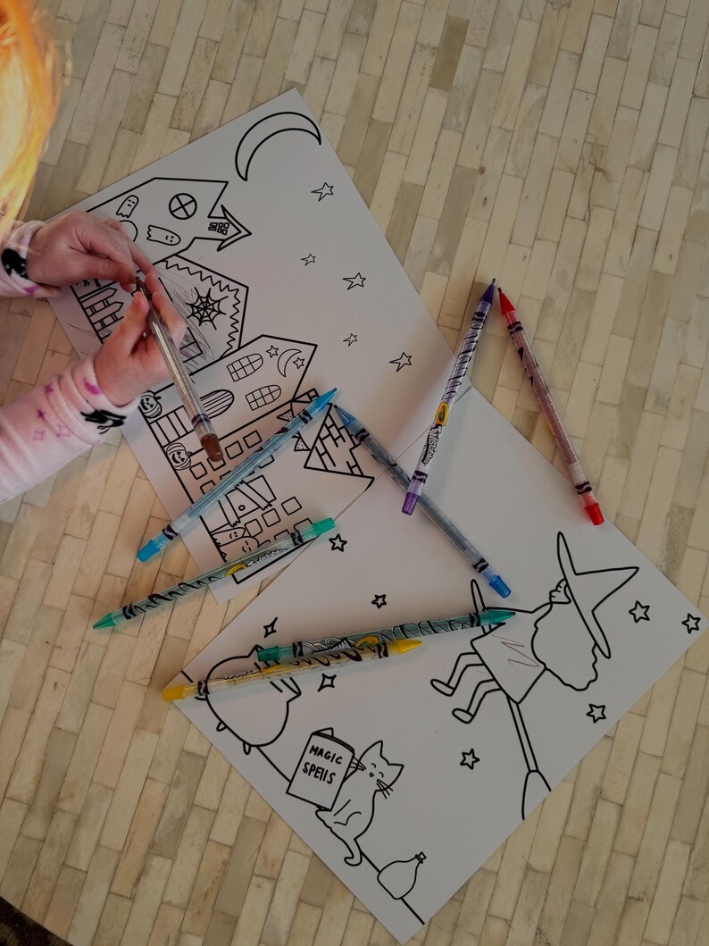 Puede incluir: Papel blanco con dibujos lineales negros de una casa embrujada, una bruja en una escoba y estrellas. Varios l&aacute;pices de colores est&aacute;n esparcidos alrededor de los dibujos. Una persona sostiene un bol&iacute;grafo sobre el dibujo.