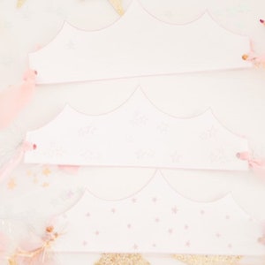 May include: Three white paper crowns with pink star designs. The crowns are on a white surface with pink and gold glitter.