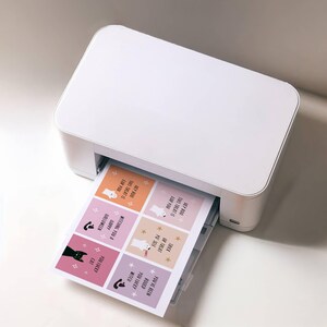 May include: A white printer printing a sheet of Halloween-themed stickers. The stickers feature cartoon ghosts, cats, and text like "You Lucky Witch" and "Hey Boo, This Treat is For You".