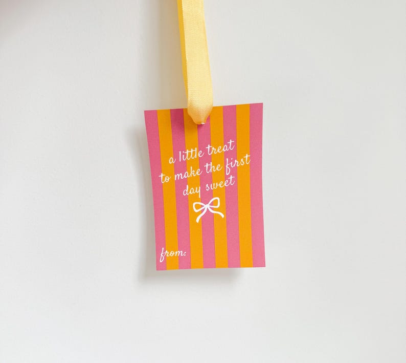 Pu&ograve; includere: Un'etichetta regalo rettangolare con strisce verticali rosa e arancioni, sospesa da un nastro giallo. L'etichetta presenta il testo "a little treat to make the first day sweet" e un'immagine di fiocco.