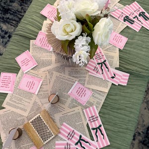 Pode incluir: Uma mesa decorada com uma toalha de mesa verde, p&aacute;ginas de livros espalhadas e um arranjo floral de rosas brancas. Marcadores listrados rosa e branco com la&ccedil;os verdes e cart&otilde;es rosa com texto tamb&eacute;m est&atilde;o presentes, criando um tema de clube do livro.