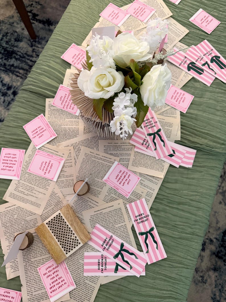 Pode incluir: Uma mesa decorada com uma toalha de mesa verde, p&aacute;ginas de livros espalhadas e um arranjo floral de rosas brancas. Marcadores listrados rosa e branco com la&ccedil;os verdes e cart&otilde;es rosa com texto tamb&eacute;m est&atilde;o presentes, criando um tema de clube do livro.