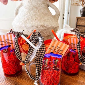 May include: Red glass mugs filled with candy and red paper shred. Each mug has a straw, a black and white checkered ribbon, and a small card that reads "BOO! HERE'S A TREAT FOR YOU HAPPY HALLOWEEN".