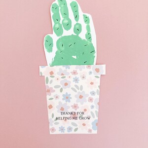 Close-up of a green handprint resembling a cactus, complete with drawn-on cactus needles, set in a pastel floral pot. Printed on sturdy white cardstock for a handmade keepsake gift