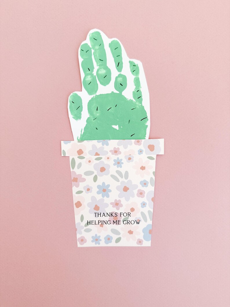 Close-up of a green handprint resembling a cactus, complete with drawn-on cactus needles, set in a pastel floral pot. Printed on sturdy white cardstock for a handmade keepsake gift