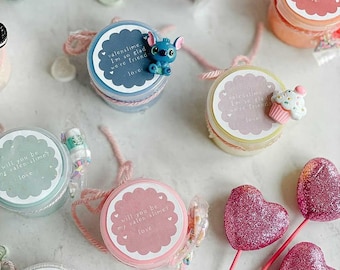 May include: A collection of small jars filled with colorful slime, each with a different Valentine's Day themed label. The labels include phrases like "Will you be my valen-slime?" and "Valenslime. I'm so glad we're friends."  The jars are decorated with pink and white string and glitter hearts.