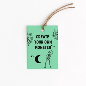 Puede incluir: Una etiqueta verde menta con el texto "CREATE YOUR OWN MONSTER" en negro. La etiqueta presenta un esqueleto negro, telara&ntilde;as, una luna creciente y estrellas. Una cuerda marr&oacute;n est&aacute; unida en la parte superior.