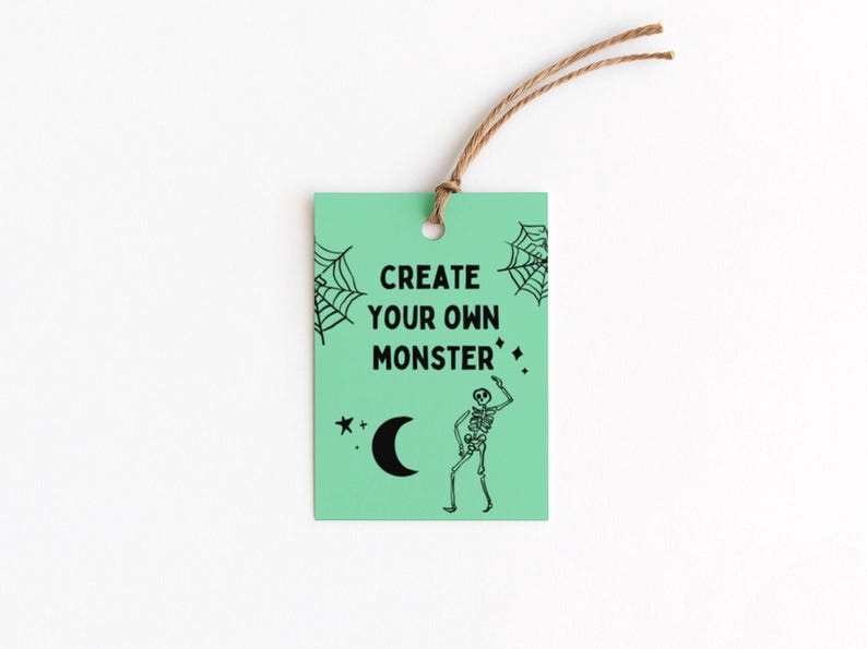 Puede incluir: Una etiqueta verde menta con el texto "CREATE YOUR OWN MONSTER" en negro. La etiqueta presenta un esqueleto negro, telara&ntilde;as, una luna creciente y estrellas. Una cuerda marr&oacute;n est&aacute; unida en la parte superior.