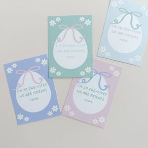 K&ouml;nnte beinhalten: Vier pastellfarbene Ostergeschenkanh&auml;nger mit einem wei&szlig;en Ei-Design und einem rosa Band. Jeder Anh&auml;nger hat den Text "I'm so egg-cited we are friends" und einen Platz zum Schreiben von "from".