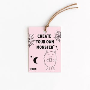 Puede incluir: Una etiqueta de regalo rosa con el texto "CREATE YOUR OWN MONSTER" en negro. La etiqueta presenta un monstruo de dibujos animados, telara&ntilde;as, una luna creciente y estrellas. Una cuerda marr&oacute;n est&aacute; atada en la parte superior.