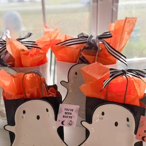May include: Halloween-themed gift sets with ghost-shaped plates, black gift boxes, and orange tissue paper. Each gift is tied with a black and white striped ribbon. The text on the card reads "YOU'VE BEEN BOOED".