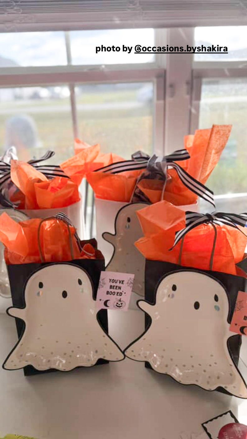 May include: Halloween-themed gift sets with ghost-shaped plates, black gift boxes, and orange tissue paper. Each gift is tied with a black and white striped ribbon. The text on the card reads "YOU'VE BEEN BOOED".